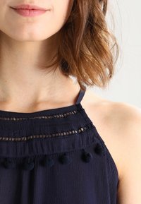 Mörkblå ärmlös topp i en strukturerad tyg, med ett scalloped halsband, dekorativa utskärningar och tofsar längs fållen.