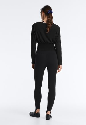 Kobieta z długimi włosami związanymi z tyłu, ubrana w czarny top z długim rękawem, legginsy z wysokim stanem oraz czarne płaskie buty, stojąca tyłem na białym tle.