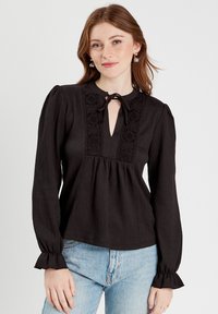 Blouse noire en tricot avec un décolleté en trou de serrure, des détails en dentelle, des manches longues avec des poignets volants, et une coupe ample. Portée avec un jean bleu clair.