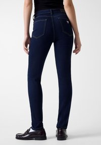 Σκούρα μπλε στενά τζιν με ομαλή υφή, διαθέτουν εφαρμοστή σχεδίαση, πίσω τσέπες και αντίθετες κίτρινες ραφές. Φοριούνται με σκούρα μοκασίνια.