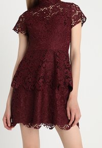 Spetsklänning i mörk burgundy med hög krage, korta ärmar och scallopad nederkant. Har blommönster och lagerpelsdesign.