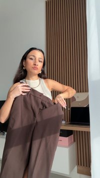 Pantalon marron tenu par une personne, présentant une coupe sur mesure, une texture lisse et des poches avant. L'arrière-plan comprend un panneau en bois et un bureau.