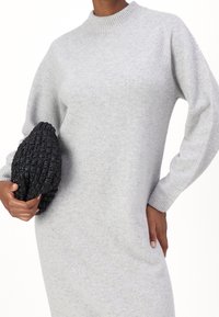 Robe en tricot gris clair avec un col montant et des manches longues. Présente une silhouette ajustée et une texture douce. Associée à une pochette tissée noire.
