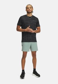 Mustat kamouflagekuosiset urheilupaita yhdistettynä vaaleanvihreisiin shortseihin, joissa on Under Armour -logo. Malli seisoo itsevarmana urheiluvaatteet jalassa.