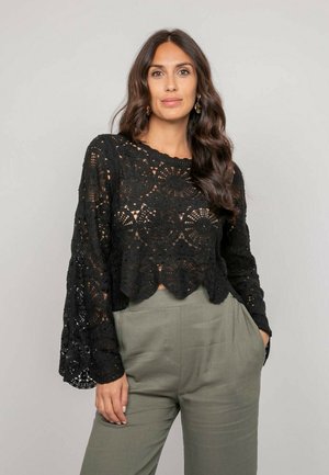 Femme aux longs cheveux ondulés portant des boucles d'oreilles dorées, un haut noir en crochet à manches longues, et un pantalon taille haute vert olive, les mains dans les poches.