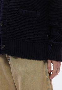 Sötétkék kötött kardigán struktúrált mintával, bordázott mandzsettákkal és szegéllyel, egyetlen zsebbel és gombos rögzítéssel. Világos nadrág fölött viselve.