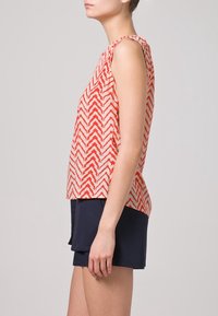 Top senza maniche con motivo a zigzag rosso e bianco, dal taglio morbido, realizzato in tessuto leggero. Abbinato a pantaloni corti scuri, texture liscia.