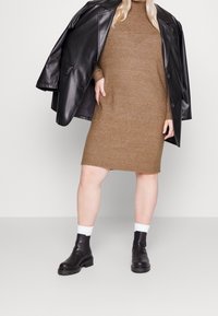 Robe-pull marron en maille avec col côtelé, associée à une veste en cuir noire oversize et des bottines noires à semelle épaisse.