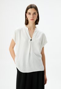 Blusa bianca a maniche corte con scollo a V e dettaglio di bottoni neri al collo, realizzata in un tessuto liscio e leggero. Design morbido e fluido.