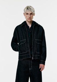 Ung person med kort blond hår iført en sort denimjakke og matchende vide bukser mod en simpel hvid baggrund.