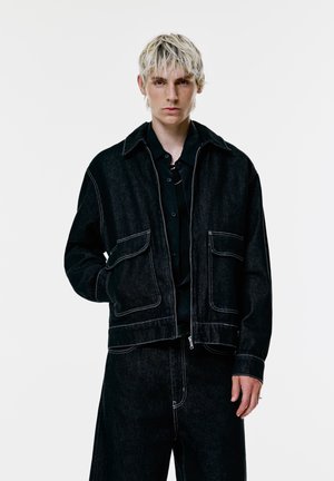 Jongere met kort blond haar die een zwart denim jack en bijpassende wijde broek draagt tegen een eenvoudige witte achtergrond.