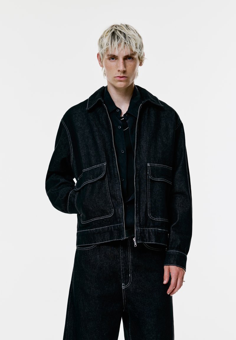 Ung person med kort blond hår iført en sort denimjakke og matchende vide bukser mod en simpel hvid baggrund.