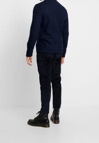 Granatowy sweter dzianinowy z wysokim dekoltem, połączony z ciemnymi spodniami sztruksowymi. Czarne buty na sznurówki z grubą podeszwą. Gładka tekstura, minimalne detale.
