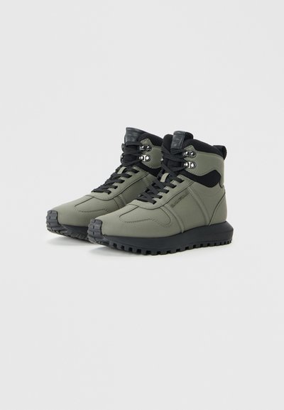 Green waterproof high-top sneakers with black accents, featuring a textured rubber sole, lacing system, and metallic eyelets.
