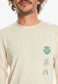 Cremfarbendes Langarmshirt mit grünen Grafikdesigns auf der Vorderseite: ein Spiralmotiv, ein Friedenssymbol und wellenförmige Linien. Weicher, leichter Stoff.
