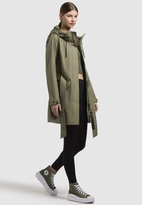 Olijfgroene waterdichte jas met een trekkoord capuchon, riem en een asymmetrische zoom, gecombineerd met zwarte leggings en olijfgroene high-top sneakers.