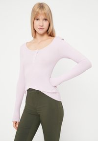 Top de manga longa em tom de roxo claro, com decote arredondado e uma nervura, apresentando uma lista de botões, combinado com calças justas de tom verde escuro.