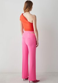 Frau mit langen Haaren, die ein asymmetrisches oranges Oberteil und weite, hochtaillierte rosa Hosen trägt, steht vor einem schlichten hellen Hintergrund.