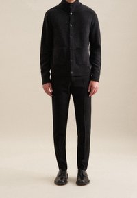 Tummanharmaa villainen neuletakki, jossa on korkea kaulus, neljä nappia ja kaksi etutaskua, yhdistetty mustiin housuihin ja nahkakenkiin.