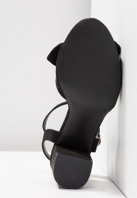 Sandale noire pour femme avec talon bloc, semelle visible et bride à la cheville, positionnée à la verticale contre une surface blanche.