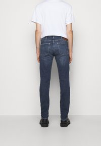 Man som bär mörkblå slim-fit jeans, vit t-shirt och svarta skor står vänd mot en enkel vit vägg från baksidan.