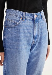 Ljusblå denimjeans har hög midja, klassisk femfickdesign, kopparnitar och subtila urtvättningar med en texturerad yta.