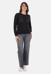 Maglione nero a maglia con un motivo a diamante aperto, scollatura rotonda e maniche lunghe, abbinato a jeans grigi a gamba larga e sneakers bianche.