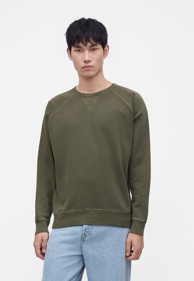 Jeune homme portant un sweat-shirt vert olive uni et un jean bleu clair, debout devant un fond blanc.