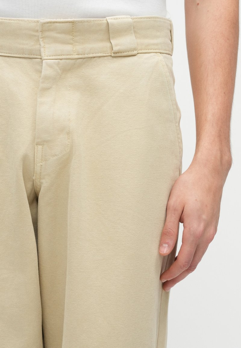 Pantaloni in cotone beige con superficie texturizzata, dotati di cintura e tasche laterali, che evidenziano una vestibilità rilassata e linee pulite.