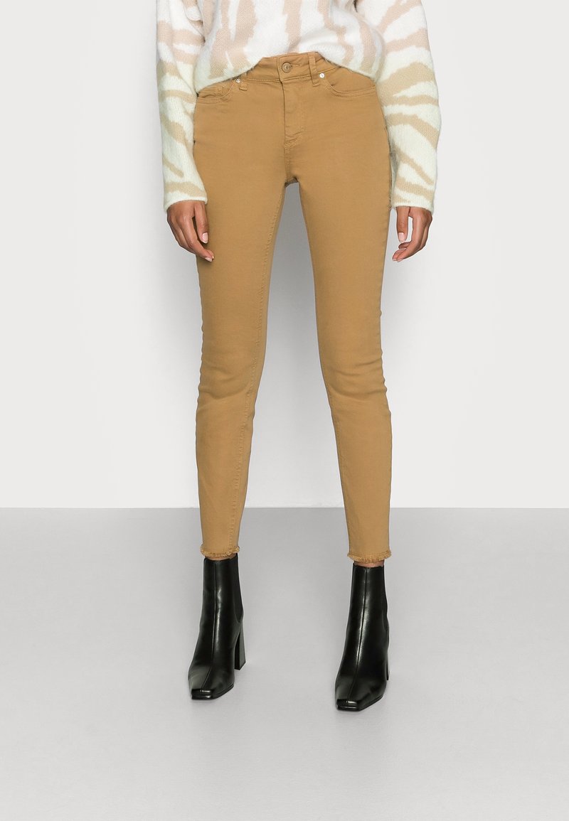 Femme portant un pantalon skinny camel taille haute avec des ourlets effilochés, un pull beige et blanc rayé, et des bottines à talons noires.