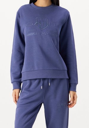 Kobieta w niebieskiej bluzie i dopasowanych spodniach dresowych z wyszywanym z przodu logo "GANT American Sportswear".