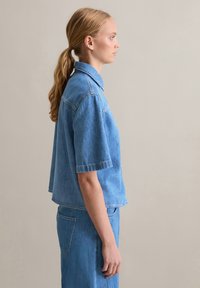 Short-sleeve denim shirt in light blue, featuring a classic collar, a relaxed fit, and visible stitching details. Textured cotton fabric.