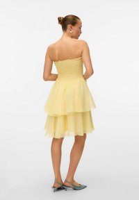 Robe jaune sans bretelles avec un corsage froncé et une jupe à superpositions ; tissu léger et transparent ; portée avec des chaussures à talons ouverts teal.