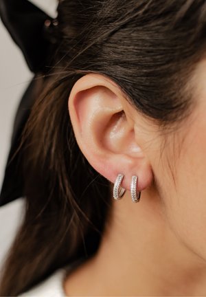 Pendientes de aro de plata con una superficie texturizada, adornados con pequeñas piedras brillantes y claras, colgando por debajo del lóbulo de la oreja.