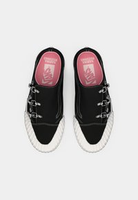 Zapatos negros sin cordones con suelas de goma blanca texturizada, herrajes plateados y plantillas rosas que presentan un logo y texto impreso.