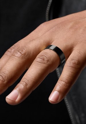 Et svartmetall-band ring på en hånd, med en jevn, matt finish og et flatt design. Naturlig hudtone og upolerte negler er synlige.