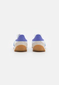 Ett par vita och blå sneakers med lila hälflikar och gummisula, sedd bakifrån på en vit bakgrund.