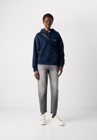 Mørkeblå hettegenser med frontlomme og snøring, paret med lysegrå jeans og hvite sneakers, stående mot en enkel bakgrunn.