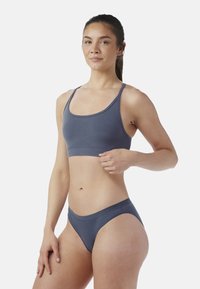 Mujer vestida con sujetador deportivo azul y ropa interior alta a juego, posando con una mano descansando en su cintura contra un fondo blanco.