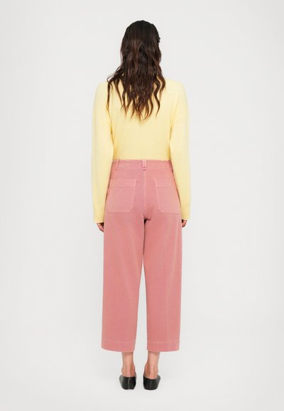 Woman with long wavy hair wears pale yellow sweater, high-waisted wide-leg pink pants, and black slip-on shoes, standing against white background.