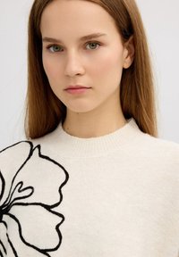 Suéter de color beige claro con un diseño de bordado floral negro, cuello redondo y textura suave, presentado sobre un fondo neutro.