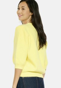 Vrouw met lang, donker haar die een gestructureerde gele trui met driekwartmouwen en donkerblauwe jeans draagt, glimlachend over haar schouder.