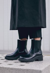 Czarne skórzane botki za kostkę z elastycznymi panelami po bokach, masywną gumową podeszwą i eleganckim designem, spoczywające na teksturowanej szarej powierzchni.