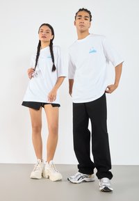 Hvite overdimensionerte t-skjorter med blå logo, kombinert med svarte shortser og bukser. Fottøy: kraftige hvite joggesko og sølvfargede sportssko.