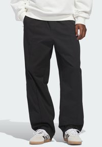 Μαύρο παντελόνι με φαρδύ πόδι από ελαφρύ ύφασμα, με πιέτες στο μπροστινό μέρος και πλαϊνές τσέπες, συνδυασμένο με λευκά αθλητικά παπούτσια με μαύρες ρίγες.