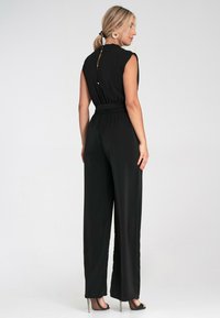 Czarny kombinezon z dopasowaną talią, krótkimi rękawami i detalem w kształcie otworu z tyłu. Posiada szerokie nogawki oraz gładką fakturę materiału.