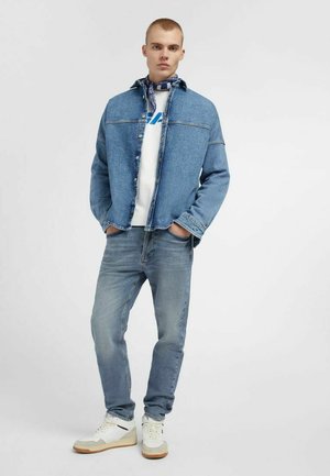 Uomo in piedi con le mani in tasca che indossa una giacca di jeans blu, una maglietta bianca con grafica, jeans blu, sneakers bianche e una sciarpa al collo blu con motivo a fantasia.