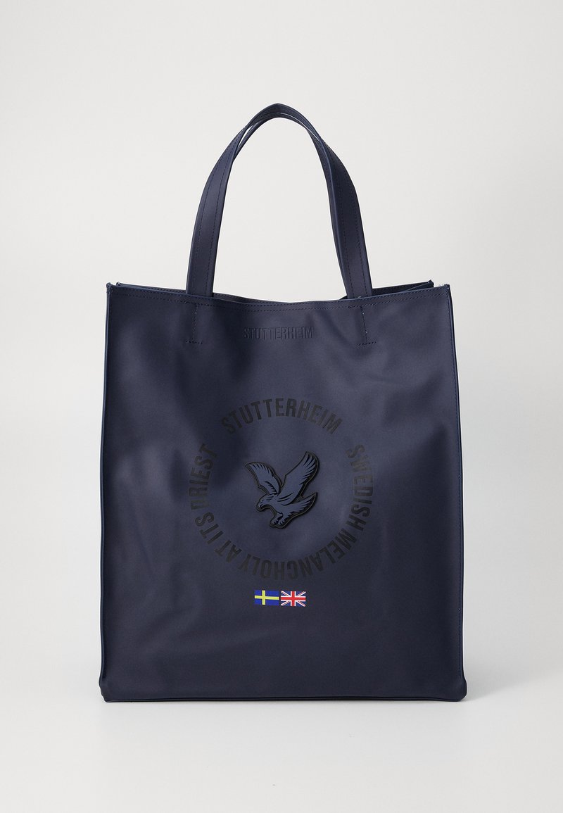 Mørkeblå tote bag laget av glatt materiale, med preget logo og fugledesign, to slanke håndtak og flaggdetaljer.