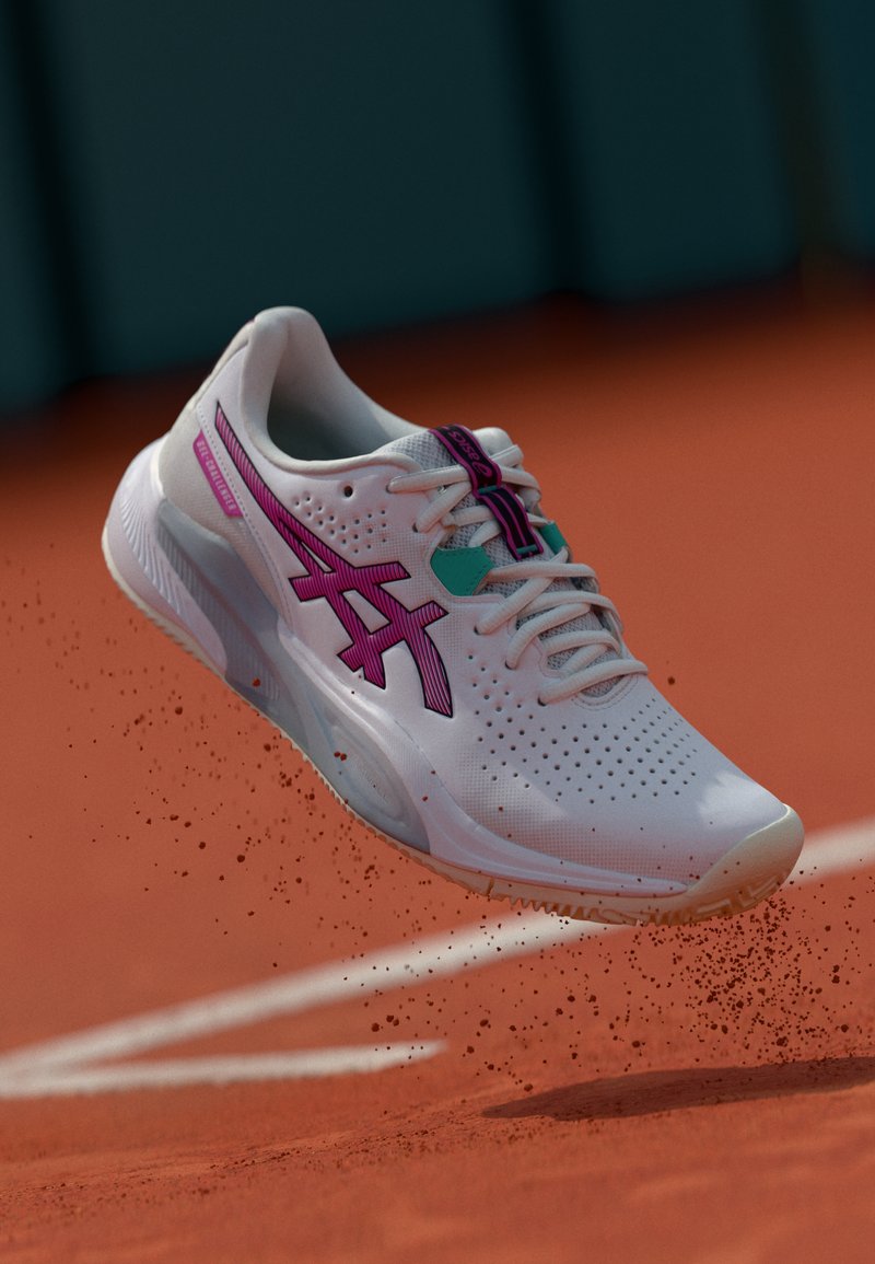 Biała sportowa sneakerka z różowymi akcentami i teksturowaną cholewką, z perforacjami, sznurówkami i gumową podeszwą, unosząca się nad powierzchnią glinianą.