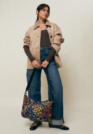 Mujer con chaqueta beige, blusa marrón, vaqueros de pierna ancha y zapatos planos negros, sosteniendo un bolso de hombro con estampado colorido sobre un fondo liso.